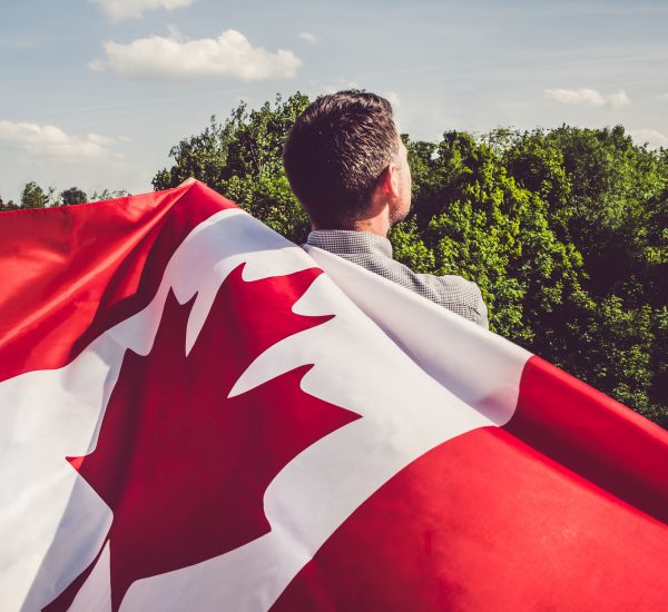 Attractive man holding Canadian Flag on blue sky background on a clear, sunny day. View from the back, close-up. National holiday concept